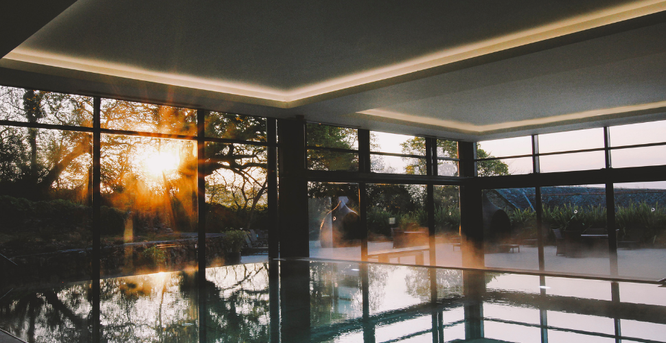 Indoor spa pool with floor-to-ceiling windows overlooking a landscaped garden, with warm sunlight streaming through the trees and reflecting across the still water. Indoor spa pool with floor-to-ceiling windows overlooking a landscaped garden, with warm sunlight streaming through the trees and reflecting across the still water.